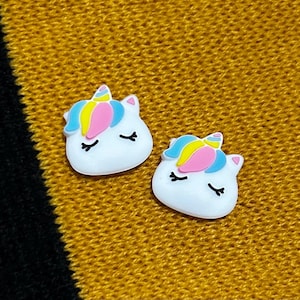 May include: Two white unicorn-shaped charms with pink, blue and yellow details. The charms have closed eyes and a single horn.