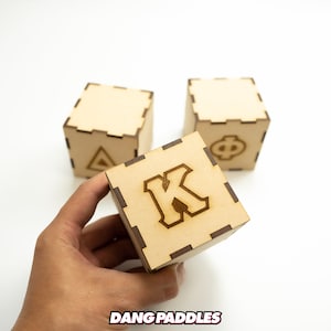 May include: Three wooden blocks with letters A, K, and Phi engraved on them. The blocks are light brown and have a slightly rough texture. The block with the letter K is being held in a hand.