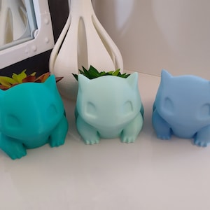 Op de afbeelding: Drie keramische potten in de vorm van een Pokémon-personage, elk in een andere kleur: teal, lichtblauw en blauw. De potten zijn gevuld met kleine groene planten.