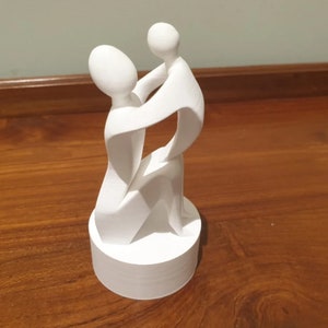 May include: White abstract sculpture of two figures embracing, one holding the other, on a white circular base.