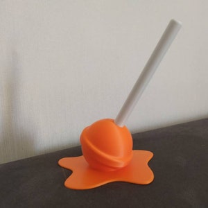 May include: Orange lollipop with a white stick, the lollipop is melting and the melted part is spreading out on a dark surface.