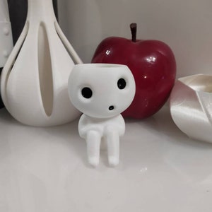 Può includere: Vaso da fiori in ceramica bianca a forma di piccola figura seduta con occhi grandi e una piccola bocca. Il vaso è seduto su una superficie bianca accanto a una mela rossa.