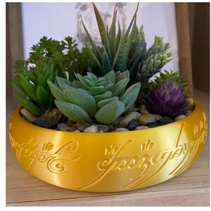 Peut inclure: Un pot de fleurs en or en forme de l'Anneau Unique du Seigneur des Anneaux, avec l'inscription "Un anneau pour les gouverner tous" gravée autour du bord. Le pot est rempli de différentes succulentes et d'une fleur violette.