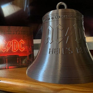 May include: A silver bell with the band "AC/DC Hell's Bell" inscribed on it. The bell is sitting on a wooden surface next to a CD case with the band's logo on it.