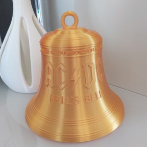 May include: A gold-colored bell with the words "AC/DC Hell's Bell" inscribed on it. The bell is made of plastic and has a smooth, shiny finish.