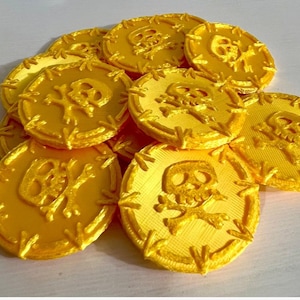 May include: A collection of yellow, 3D-printed pirate coins. Each coin features a skull and crossbones design in the center. The coins are round with a textured edge, and are scattered across a white surface.