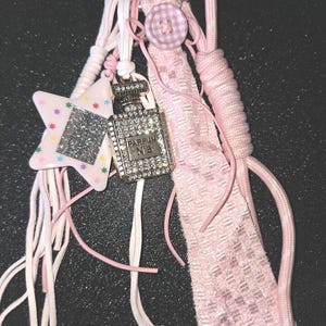 May include: A pink and white accessory with a star-shaped charm, a rhinestone-studded perfume bottle charm, and a pink fabric element. The perfume bottle charm has the text "PARFUM N°5". The accessory is attached to a metal clasp.
