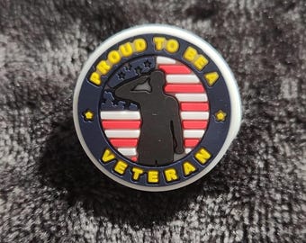 May include: A blue and white round charm with the text "Proud to be a Veteran" in yellow lettering. The charm features a silhouette of a person saluting in front of an American flag.