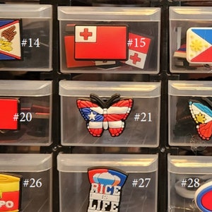 May include: A collection of colorful shoe charms in a clear plastic storage case. The charms include flags, butterflies, and other designs. The charms are numbered 14 through 28.