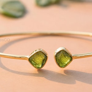 May include: A gold-coloured bracelet featuring two square, light green gemstones. The bracelet has an open design, with the gemstones at each end. The background is a light peach colour.