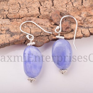 Puede incluir: Un par de pendientes con piedras ovaladas de color lavanda y detalles metálicos plateados. Los pendientes tienen un diseño de gancho para orejas perforadas. El fondo incluye un trozo de madera.