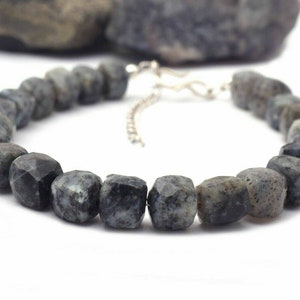 May include: A necklace featuring faceted labradorite beads in shades of grey and blue-grey. The beads are strung on a silver-coloured chain with a matching clasp. The necklace is presented against a white backdrop.