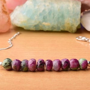 Könnte beinhalten: Ein silbernes Kettenarmband mit einer Reihe facettierter, ovaler Rubinperlen mit grünen Einschlüssen. Das Armband wird auf einer hellbraunen Oberfläche präsentiert, mit farbigen Edelsteinen im Hintergrund.