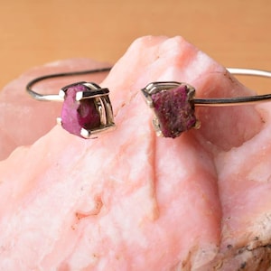 May include: A silver-tone cuff bracelet featuring two raw, rough-cut ruby gemstones. The rubies are a deep pinkish-red color and are set in square, open-frame settings. The bracelet is displayed on a pink stone surface.