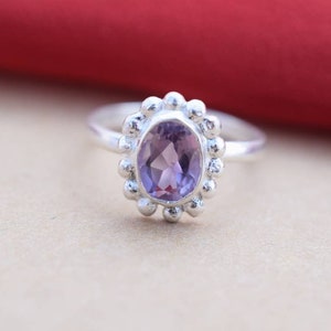 May include: A silver ring featuring an oval, light purple gemstone. The gemstone is surrounded by a beaded, floral-like design. The ring is set against a neutral background with a red accent.