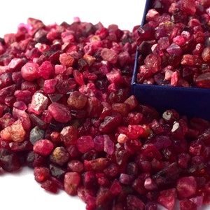May include: A close-up of a pile of red ruby gemstones. The rubies are of various sizes and shapes, and some have a slightly darker hue. The rubies are scattered on a white surface.