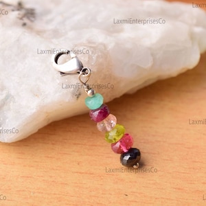 Könnte beinhalten: Ein Anhänger mit einem silbernen Verschluss und einer Kette aus bunten Edelsteinen. Zu den Edelsteinen gehören grüne, rosa, rote und schwarze Perlen. Der Anhänger wird auf einer weißen und beigen Oberfläche präsentiert.