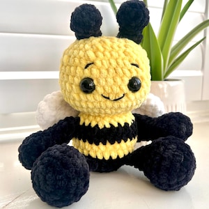 Puede incluir: Un peluche de abeja hecho a mano con ganchillo. La abeja tiene un cuerpo amarillo con rayas negras, antenas negras y ojos grandes negros. Tiene alas blancas y patas negras. El peluche está sobre una superficie blanca.