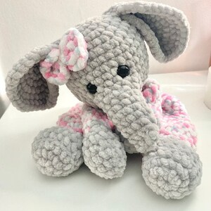 May include: A handmade grey crochet elephant with large ears, black eyes, and a pink and white bow. The elephant has a pink and white scarf and is made of soft, textured yarn. A cute, cuddly stuffed animal.