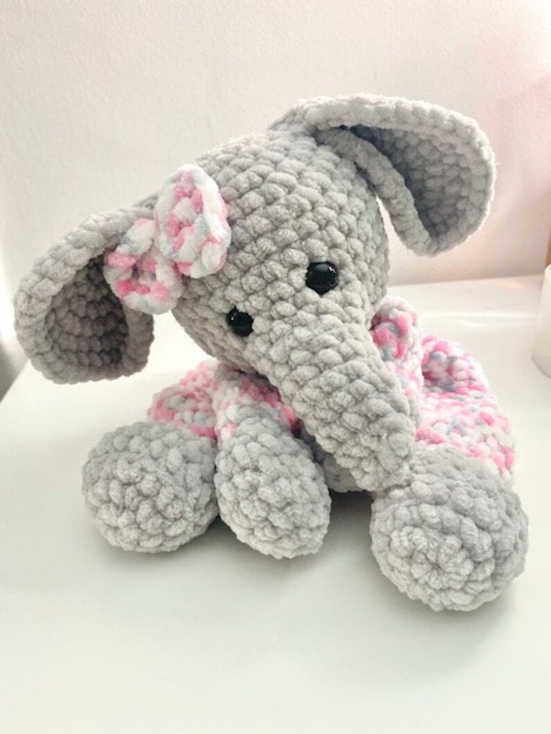 May include: A handmade grey crochet elephant with large ears, black eyes, and a pink and white bow. The elephant has a pink and white scarf and is made of soft, textured yarn. A cute, cuddly stuffed animal.