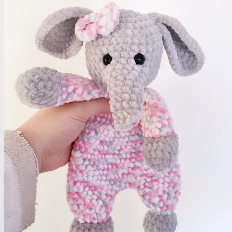 May include: A grey crocheted elephant doll wearing a pink and white patterned onesie and a matching bow. The elephant has large grey ears, a long trunk, and black button eyes. The doll is soft and plush.
