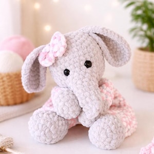 May include: A handmade grey crochet elephant with a pink bow and a pink and white patterned blanket. The elephant has black button eyes and large, floppy ears. The plush toy is soft and cuddly, ideal for a baby or child.