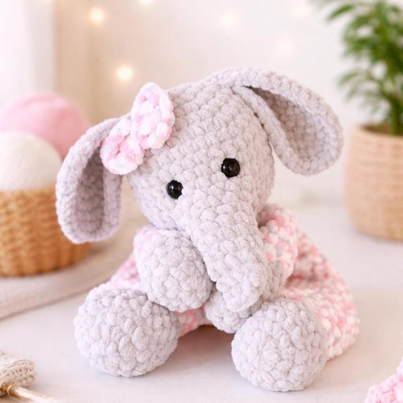 May include: A handmade grey crochet elephant with a pink bow and a pink and white patterned blanket. The elephant has black button eyes and large, floppy ears. The plush toy is soft and cuddly, ideal for a baby or child.
