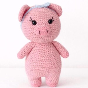 May include: A handmade pink crochet pig doll with a light blue headband. The pig has black eyes, eyelashes, and a small snout. The doll is standing upright with its arms at its sides. This soft toy is perfect for children.