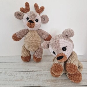 May include: Two crocheted stuffed animals, a deer and a bear, are sitting on a white wooden surface. The deer is brown and beige with a long snout and large ears. The bear is gray and beige with a short snout and small ears.