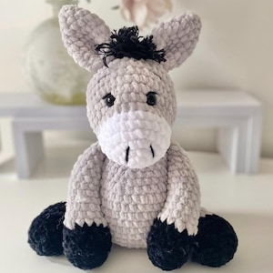 May include: A crocheted donkey toy in light grey, with a white muzzle and black hooves and mane. The soft toy has black button eyes and is sitting. A handmade item.