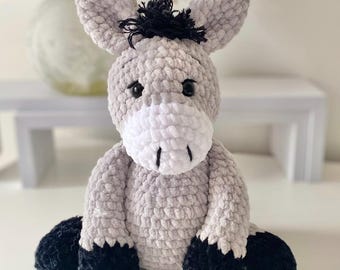 Puede incluir: Un peluche de burro hecho a mano con ganchillo. El burro es principalmente de color gris claro con un hocico blanco y pezuñas y crin negras. El animal de peluche tiene ojos negros de botón y está sentado.