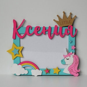 May include: A light blue picture frame with a pink unicorn, rainbow, and stars. The frame has the name "Ksenia" written in pink letters and a crown with the word "LITTLE" on it.