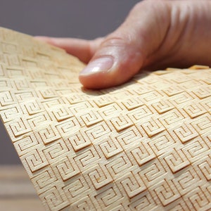 Puede incluir: Un primer plano de una fina lámina de madera flexible con un patrón geométrico tallado. La madera es de color marrón claro y el patrón está formado por pequeños cuadrados.