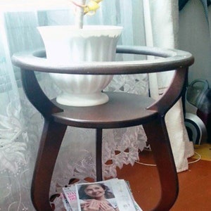 May include: A brown wooden side table with a glass top and two shelves. A white ceramic planter with a plant is on the top shelf. A magazine is on the bottom shelf.