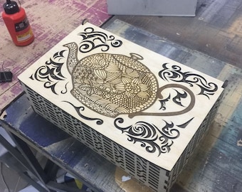 May include: A wooden box with a laser-cut design of a teapot with intricate floral patterns. The box is made of light brown wood and has a grid pattern on the bottom.