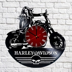 May include: Black vinyl record clock featuring a silhouette of a woman riding a Harley-Davidson motorcycle. The clock face is red with white Roman numeral hour markers. The record has the text "Harley-Davidson" and a skull logo.