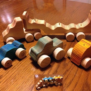 May include: A set of five wooden toy cars, including a blue car, a green car, a yellow car, and a wooden truck with a flatbed. The truck has a wooden ramp that can be lowered to load the cars.