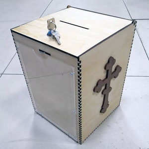 Könnte beinhalten: Eine hölzerne Spendenbox mit einer durchsichtigen Acrylglasscheibe vorne, einem Schlitz oben und einem Schlüsselloch. Die Box hat ein dekoratives Kreuzdesign an der Seite.