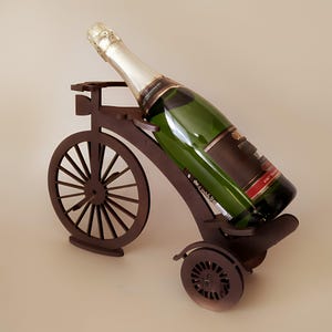 May include: A brown wooden bicycle-shaped wine bottle holder. A green wine bottle with a gold foil top is placed in the holder. The holder has a large front wheel and a smaller rear wheel.