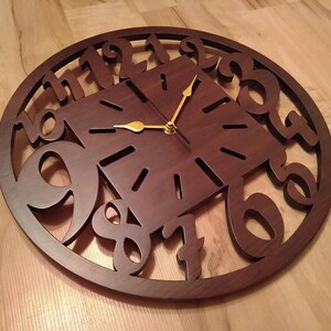 May include: A dark brown wooden wall clock with a square center and a circular outer frame. Gold-colored hands point to the time. The numbers are cut out of the wood, creating a unique design. The clock is on a wooden surface.