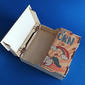 May include: A wooden box with a hinged lid. The box is decorated with a colorful cartoon unicorn, rainbow, and clouds. The text "Unicorn" is printed on the box.
