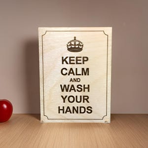 Könnte beinhalten: Ein Holzschild mit dem Text "Keep Calm and Wash Your Hands" in einer Schriftart im Vintage-Stil. Das Schild hat eine Krone über dem Text und befindet sich auf einer Holzoberfläche.
