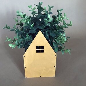 Op de afbeelding: Een houten, huisvormige plantenbak met een klein raam, gevuld met groene bladplanten.