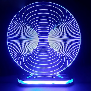 Puede incluir: Lámpara LED azul con un diseño circular con líneas concéntricas, creando una ilusión óptica 3D. La lámpara se asienta sobre una base rectangular con una luz azul brillante. El efecto general es una lámpara moderna y artística.