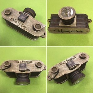May include: A wooden camera-shaped box with a glass lens and a hinged lid. The box is decorated with the words "Wedding Day" and "Memories" and has a small circle with the word "Smile" on the top.