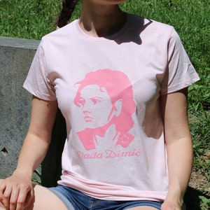 Könnte beinhalten: Eine Frau trägt ein rosa T-Shirt mit einer rosa Silhouette eines Frauenkopfes und dem Text "Nada Dimic" auf der Vorderseite. Sie sitzt auf einer Steinoberfläche mit dem Text "ZA SLOBODU 1941-1945" darauf.