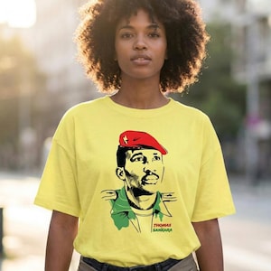 Könnte beinhalten: Ein hellgelbes T-Shirt mit kurzen Ärmeln und einer grafischen Illustration von Thomas Sankara. Das Design zeigt ein Porträt der Figur mit einer roten Baskenmütze mit Stern, vor einer stilisierten, grün-schwarzen Jacke im Militärstil.