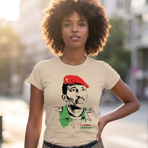 Op de afbeelding: Een beige T-shirt met korte mouwen en een grafische illustratie van Thomas Sankara met een rode baret en een groen militair uniform. Het ontwerp bevat zwarte lijntekeningen en de tekst THOMAS SANKARA in rode en groene letters.