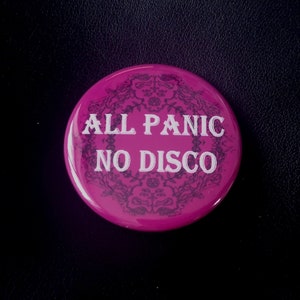Könnte beinhalten: Ein runder, magentafarbener Button mit dem weißen Text "ALL PANIC NO DISCO". Der Button hat ein dekoratives Blumenmuster in einem dunkleren Magenton. Der Hintergrund ist schwarz.