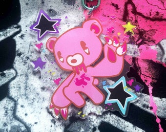 May include: Pink bear keyring with a star-shaped clasp. The bear has a distressed design with blood-like splatters and claw marks. Additional star-shaped charms in black, purple, and yellow are included. The background features a splatter design in pink, black, and white.